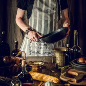 Bearded chef holding frying pan amidst rustic kitchen setup with fresh ingredients and cooking tools.