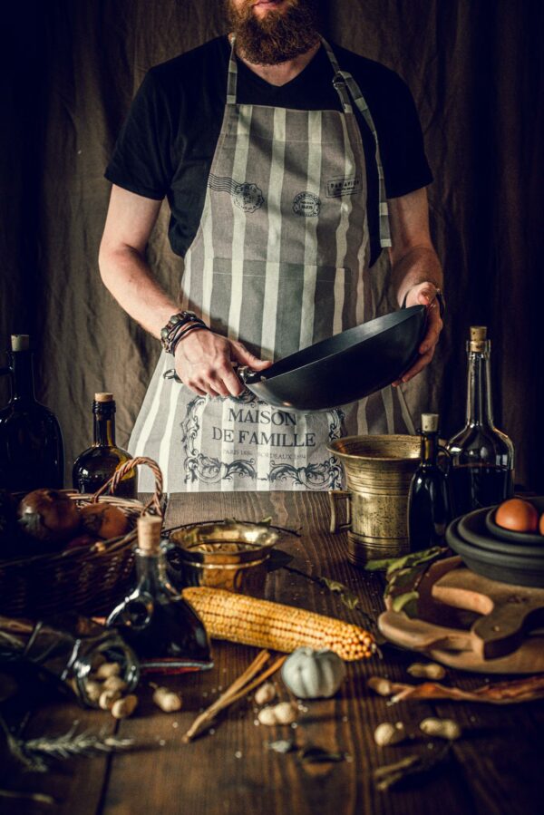 Bearded chef holding frying pan amidst rustic kitchen setup with fresh ingredients and cooking tools.