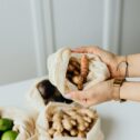 Woman's hands holding fresh turmeric in a mesh bag with ginger and limes nearby on a white table.