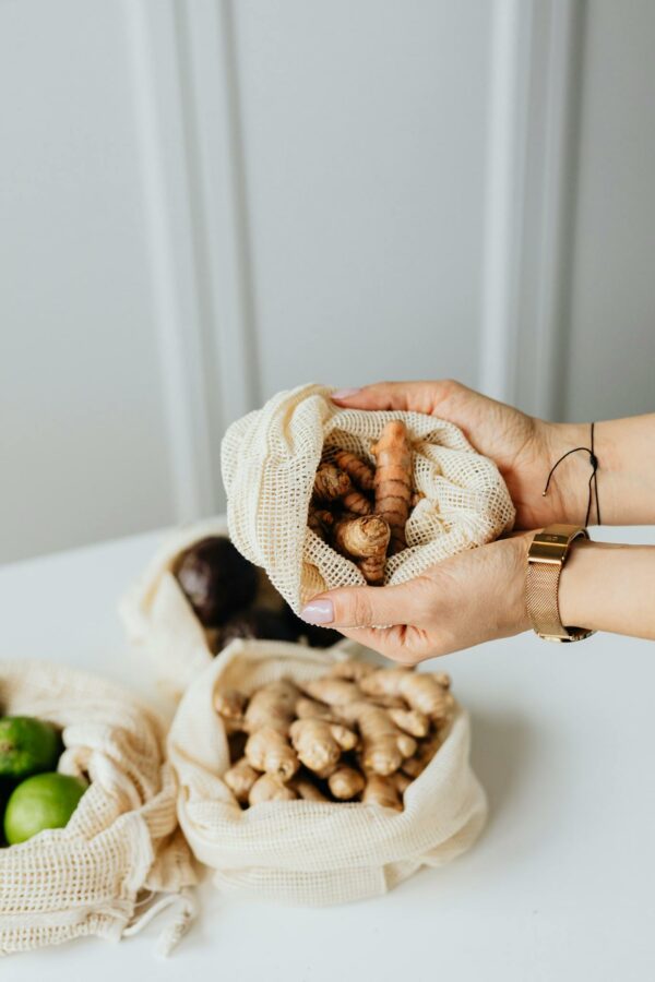 Woman's hands holding fresh turmeric in a mesh bag with ginger and limes nearby on a white table.